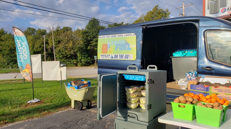 The side door of a blue van is opened, and there are bins and tables in front of it filled with food. There is a vertical fabric sign that says Food Pantry. Photo from Wesley Food Pantry Facebook page.
