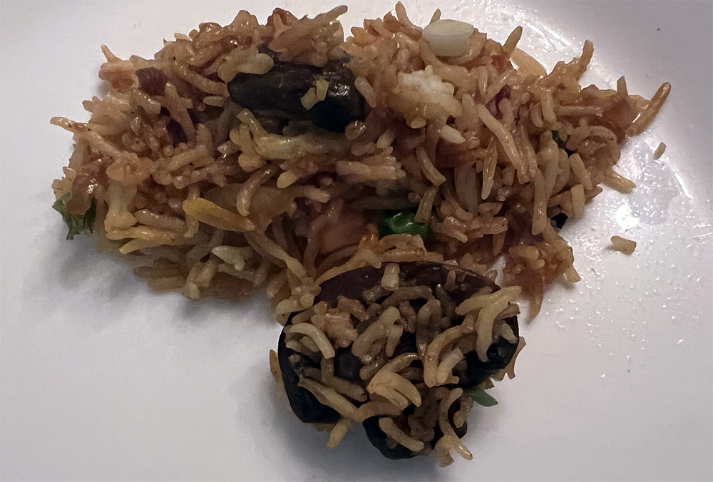 This is a photo of a small serving of mushroom fried rice on a white plate. The fried rice is a brown color with several large chunks of mushroom, a pea, and slices of green onion. Photo by Sara Ressing.