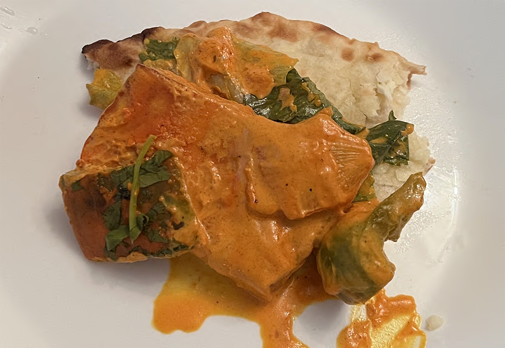 A serving of paneer tikka tandori and torn naan on a white plate. The naan bread is shades of dark and light brown and topped with a wilted bit of lettuce and a large rectange of tandoori baked paneer. There are several large chunks of green pepper and onion. All is topped with a bright orange tikka sauce and a bit of cilantro. Photo by Sara Ressing.