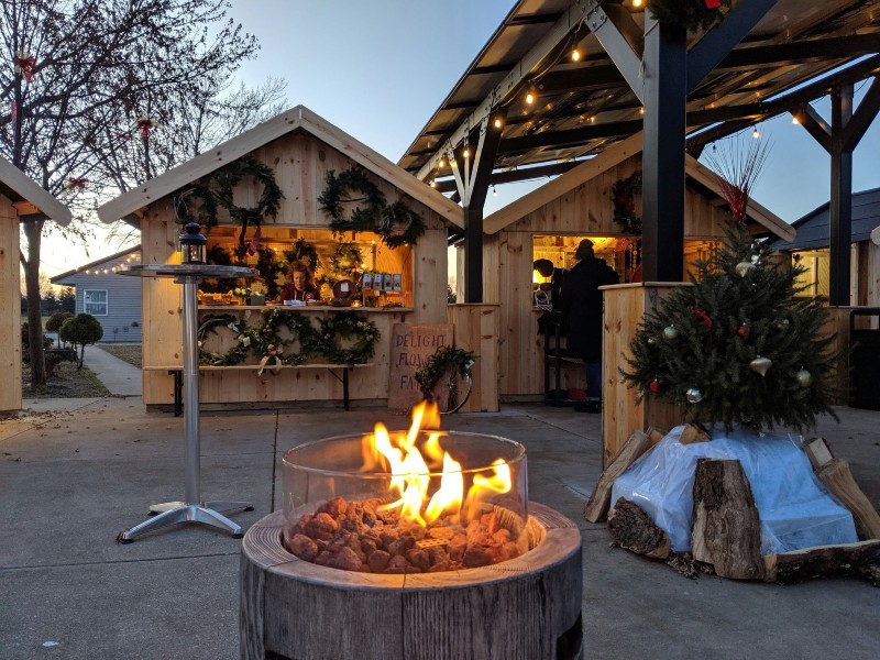 Small wooden huts with various items for sale surround the perimeter of an open area. A fire pit and high top tables are in the foreground. Photo from Riggs Christmas Market event page.