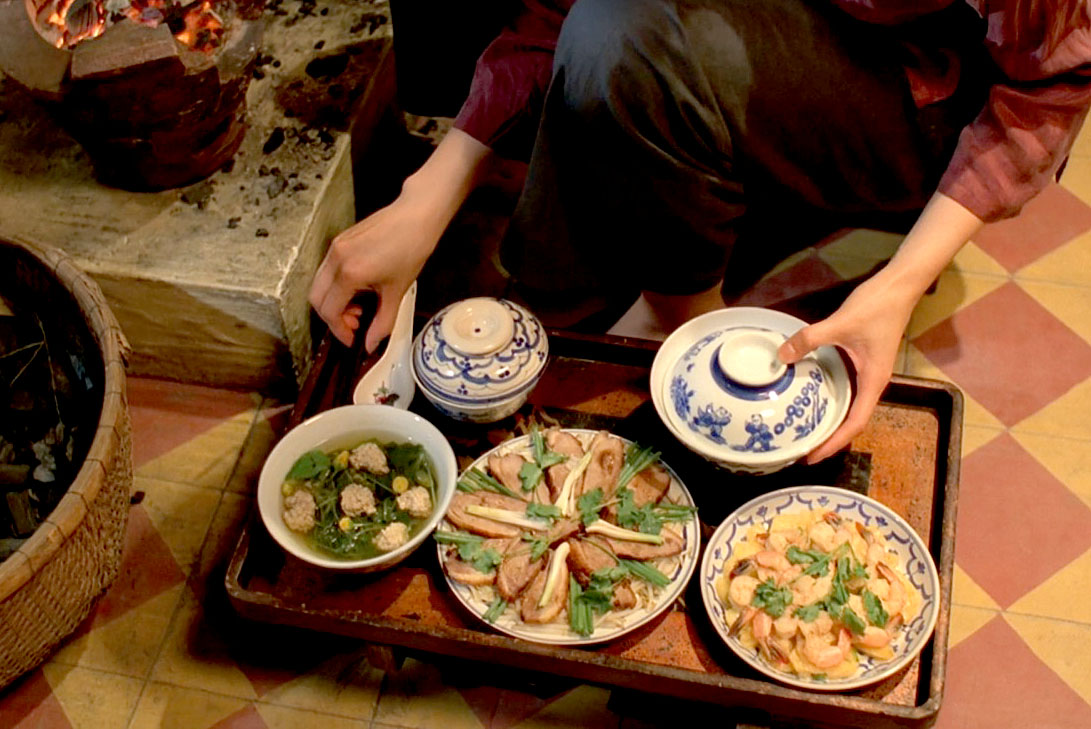 A woman with dark hair kneels before a tray of Vietnamese food in the film The Scent of Green Papaya. Photo by First Look International.