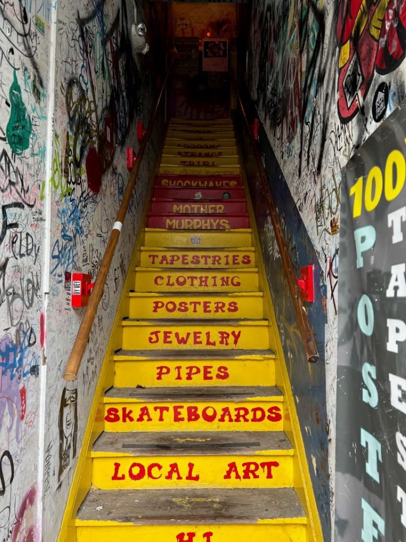 A yellow staircase in a narrow hallway. There are words painted in red on each step: Tapestries, Clothing, Posters, Jewelry, Pipes, Skateboards. The walls are covered in graffiti. Photo by Julie McClure.