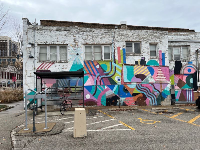 A multi-colored abstract mural painted on the side of a white brick building. A bus shelter and bike rack are in the foreground. Photo by Julie McClure.