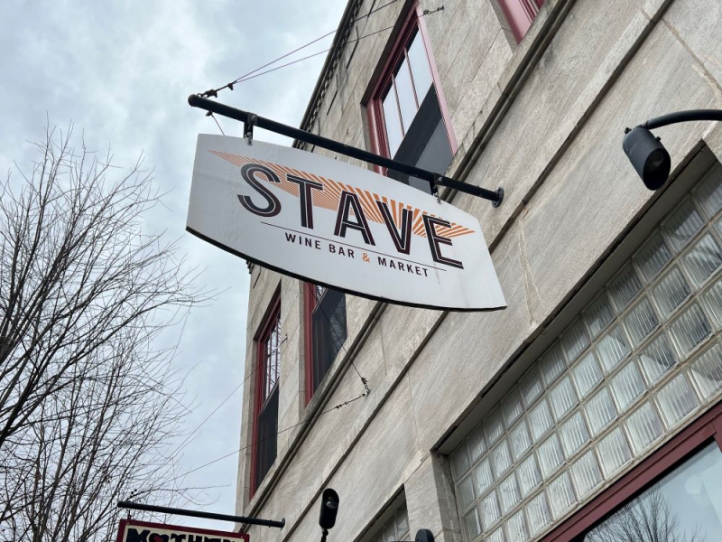 A white sign that says Stave Wine Bar & Market is hanging off the side of a gray stone building. Photo by Julie McClure.