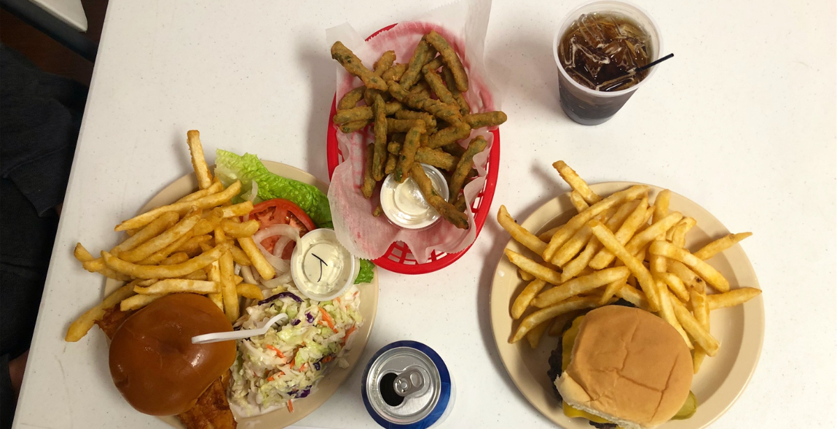 An overhead photo shows the author's lunch: a burger meal, a fish sandwich meal, and a basket of fried green beans. There is a can of Bud Light and a plastic cup with Coke. Photo by Alyssa Buckley.