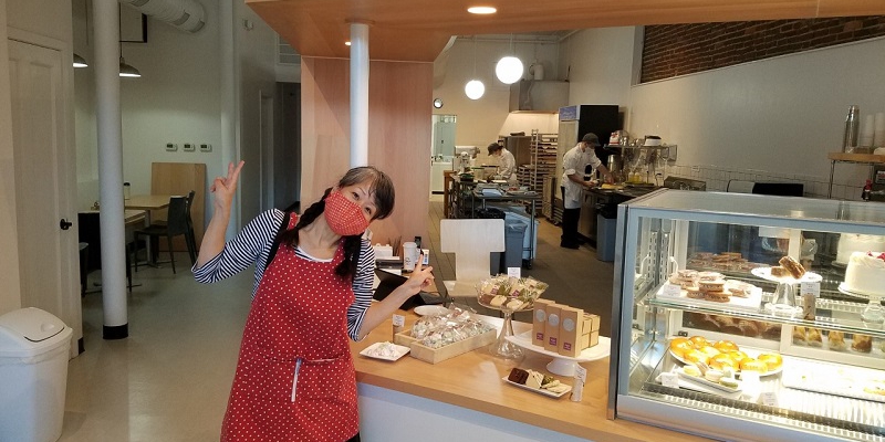 The inside of Suzu's bakery, featuring owner Jennifer Gunji, wearing a mask and apron, and holding two hands in the air with a peace sign.