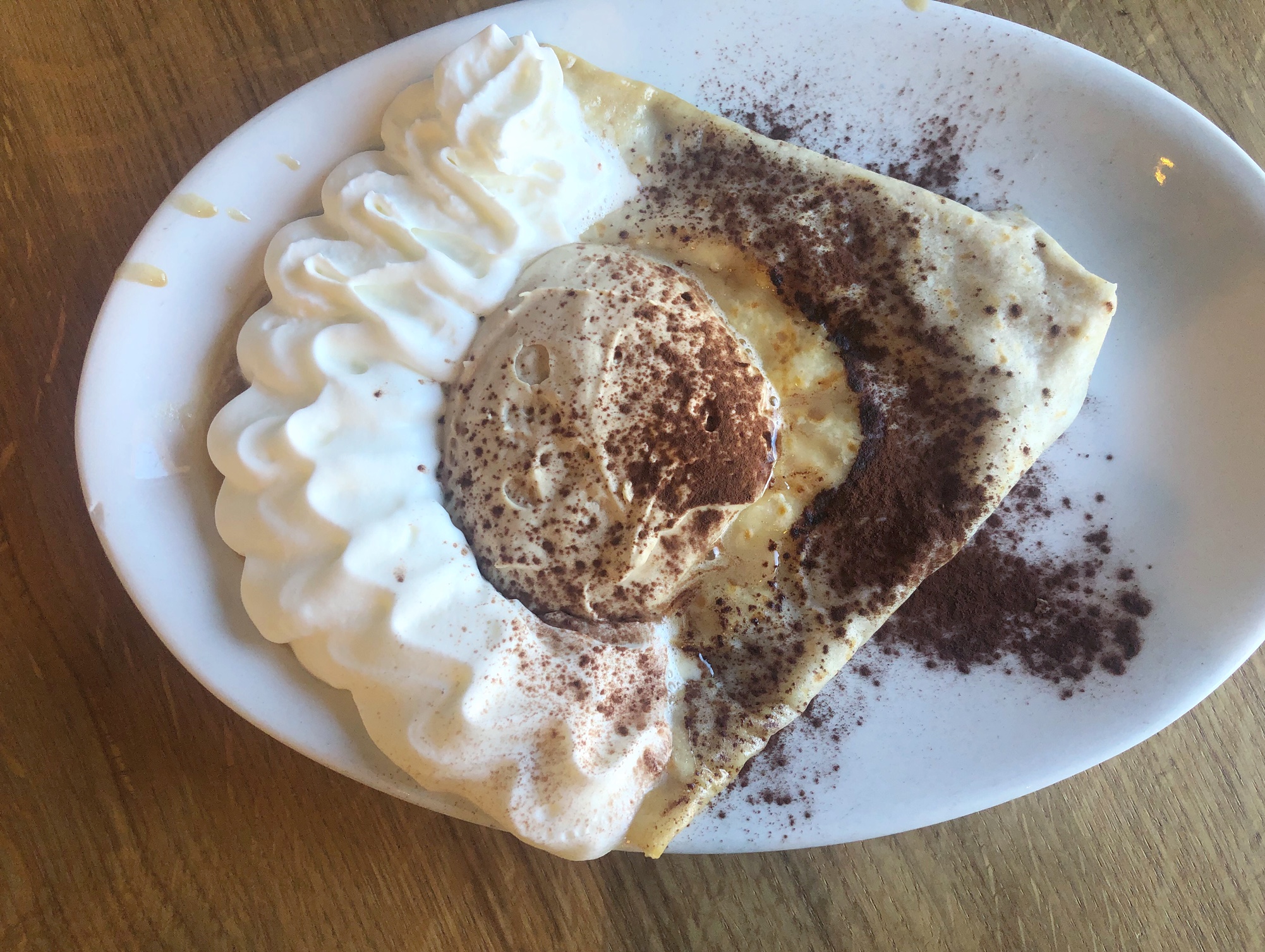 On a wooden table, there is a white plate with a tiramisu crepe on it. The crepe has whipped cream topping at the top of the triangle, a dollop of espresso whipped cream in the middle, and a dusting of cocoa powder all over. Photo by Alyssa Buckley.