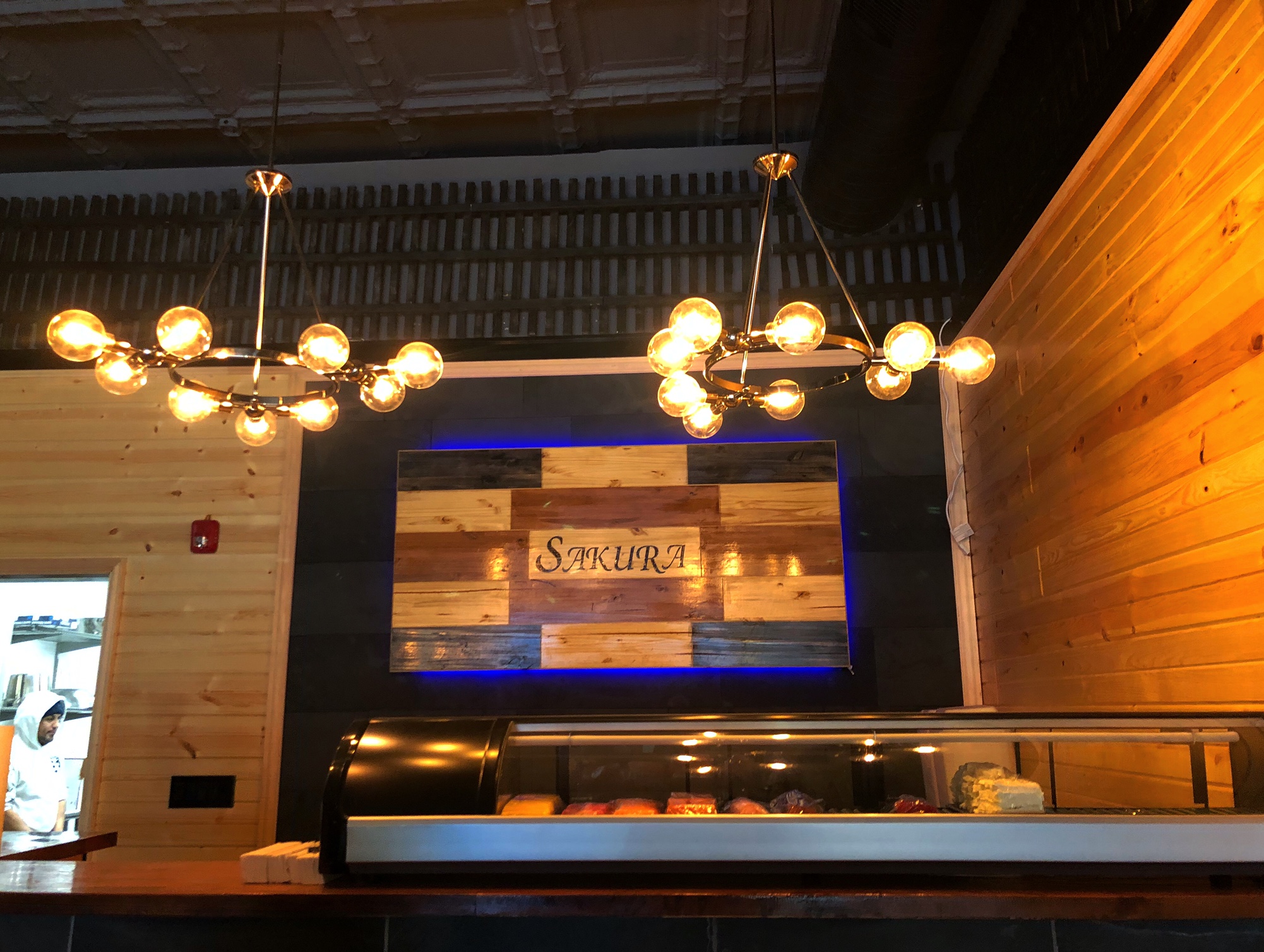 Inside Sakura, there is a sushi bar with the name of the restaurant on wood on top of a black background. There are two big beautiful golden light fixtures. Photo by Alyssa Buckley.