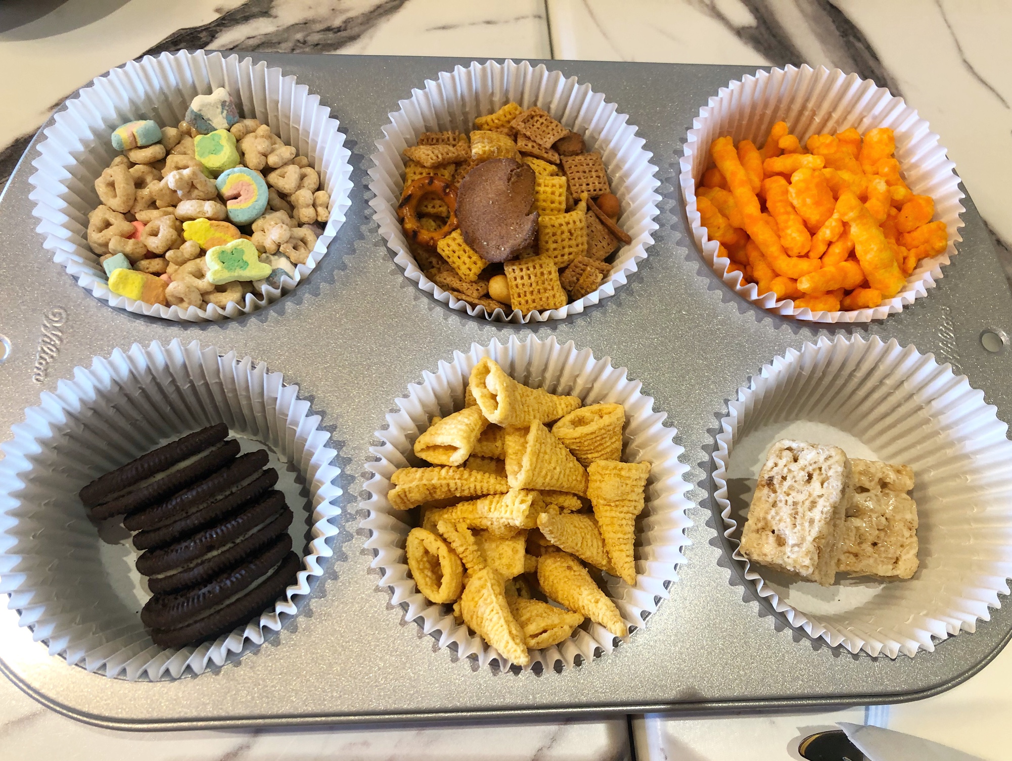 In a metal muffin tin, there are six white muffin wrappers full of processed snacks: Lucky Charms, Chex Mix, Cheetos, Oreos, Bugles, and rice krispie treats at The Literary in Downtown Champaign.  Photo by Alyssa Buckley.