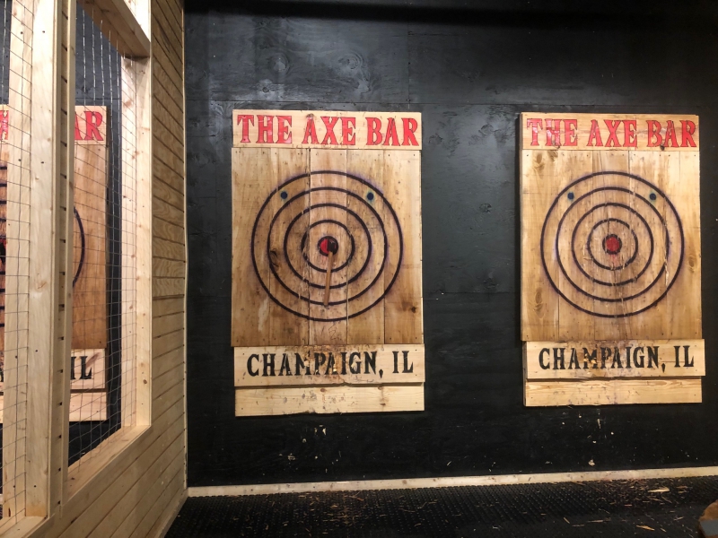 Two wooden slabs hang on a wall. They have targets with black concentric circles and say The Axe Bar in red at the top and Champaign, IL in black at the bottom. Photo by Alyssa Buckley.