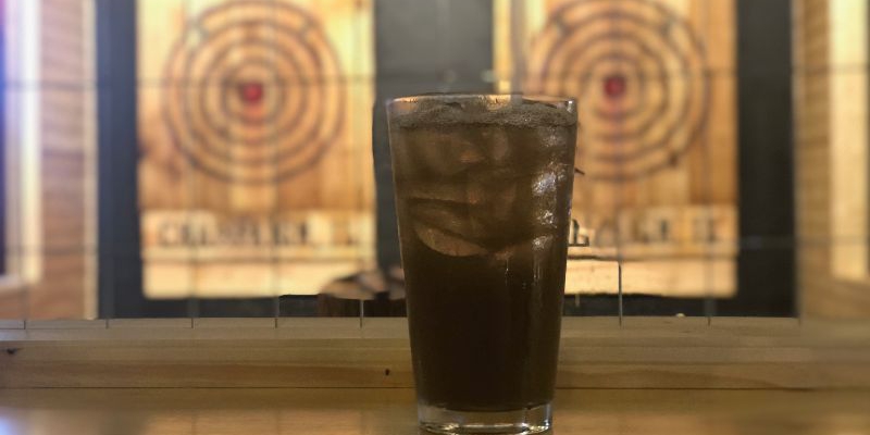 On a wooden counter, there is a dark soda-based cocktail in a highball glass. In the background are two wooden bullseye targets with a red center. Photo by Alyssa Buckley.