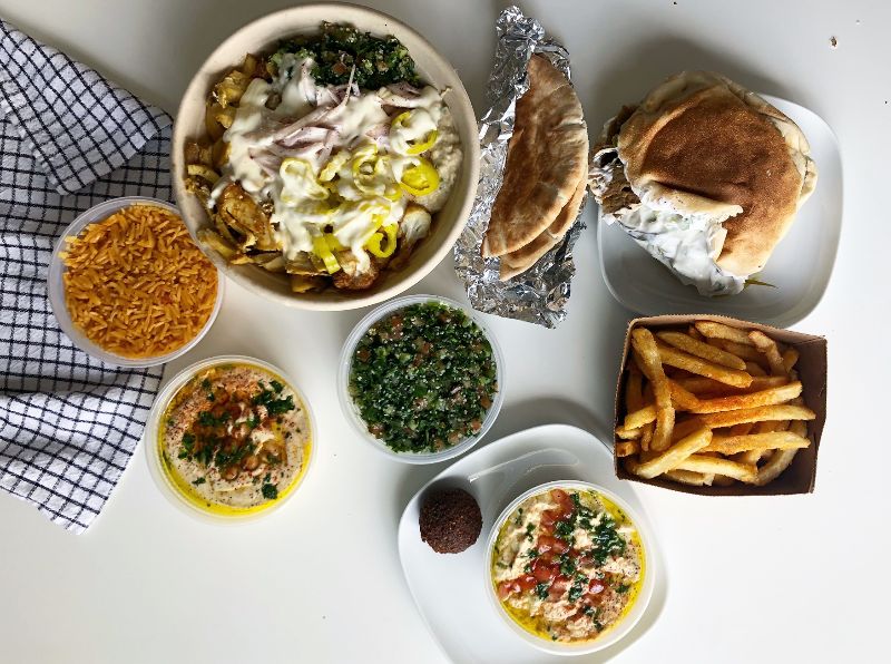 On a white table, there is a spread of Middle Eastern takeout. Two bowls of rice, one rice bowl with a white sauce, naan wrapped in tin foil, a paper basket of fries, a platter of tabbouleh, hummus, and a pita with a white sauce on a white plate. Photo by Alyssa Buckley.