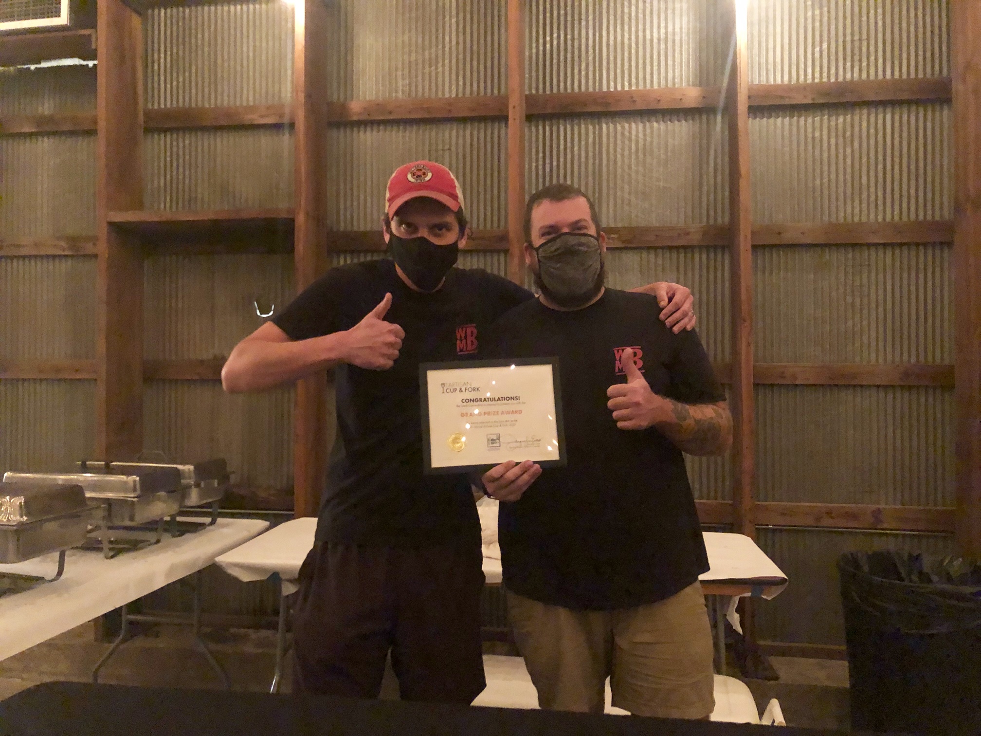 In a dimly lit barn, the duo of Weird Meat Boyz are giving thumbs up and holding the Judge's Choice Award for the Artisan Cup and Fork 2021 event. They are masked. Photo by Alyssa Buckley.