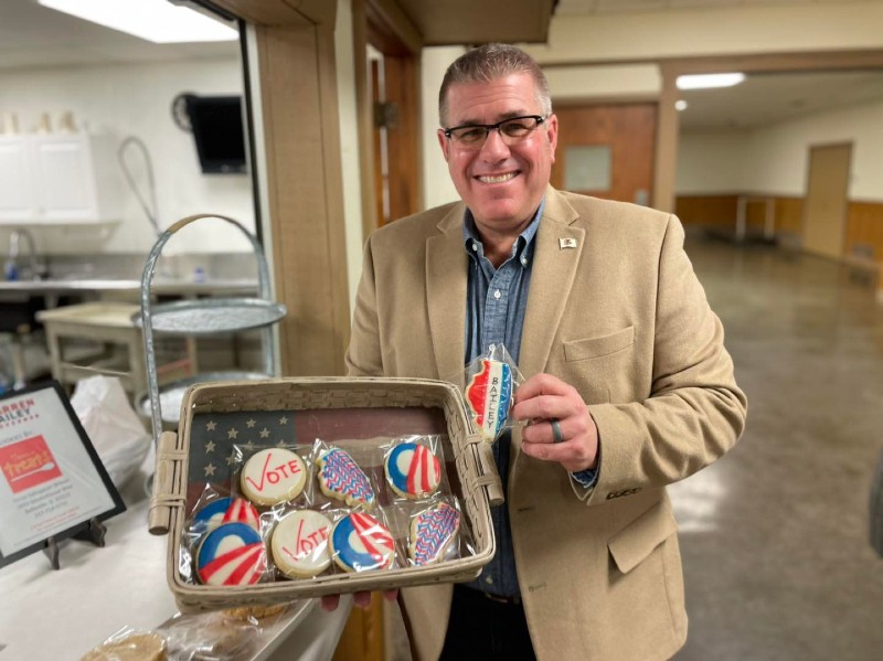 A man with short gray hair and glasses is holding a basket of cookies. Some are shaped like Illinois with white icing and red and blue designs, some are shaped like Illinois and are red, white, and blue with Bailey written down the center, some are circular with red and white waves, a blue background, and white half circle coming out of the waves. Photo from Darren Bailey for Governor Facebook page. 