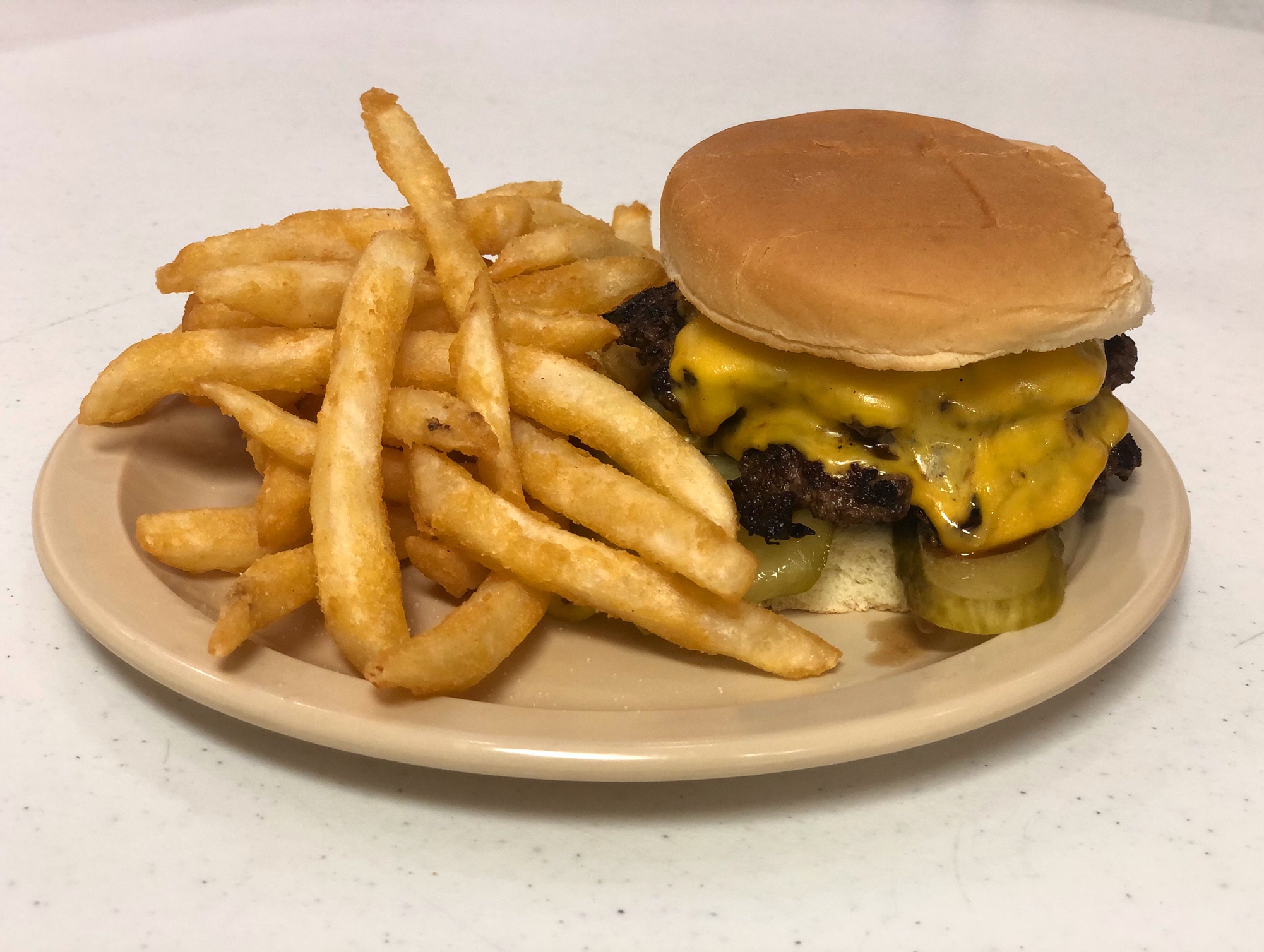 On a brown table, there is a beige plate with fries beside a cheeseburger with melty American cheese and a long pickle sticking out from under the patties. Photo by Alyssa Buckley.