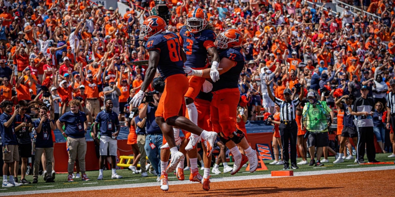Four University of Illinois football players are mid-jump toward each other on a football field. Thousands of fans are in the stands in the background. Photo from fightingillini.com. 