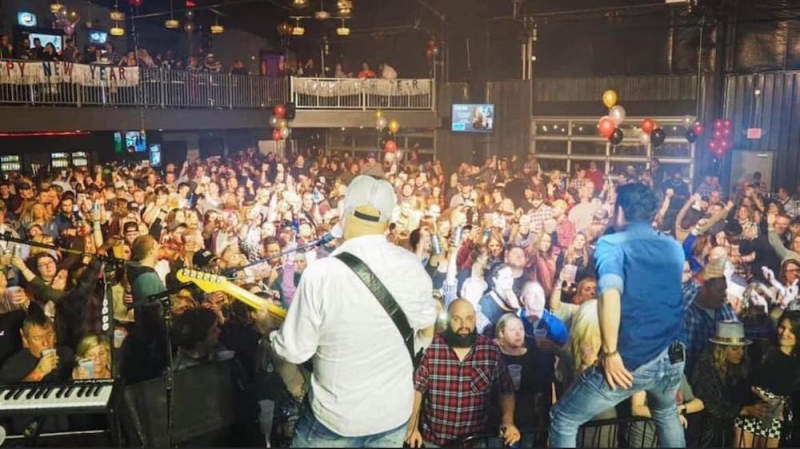 View from a stage behind band members. There is a crowd on the floor and in the balcony, with Happy New Year signs hanging from the balcony. Photo from The City Center Facebook page.