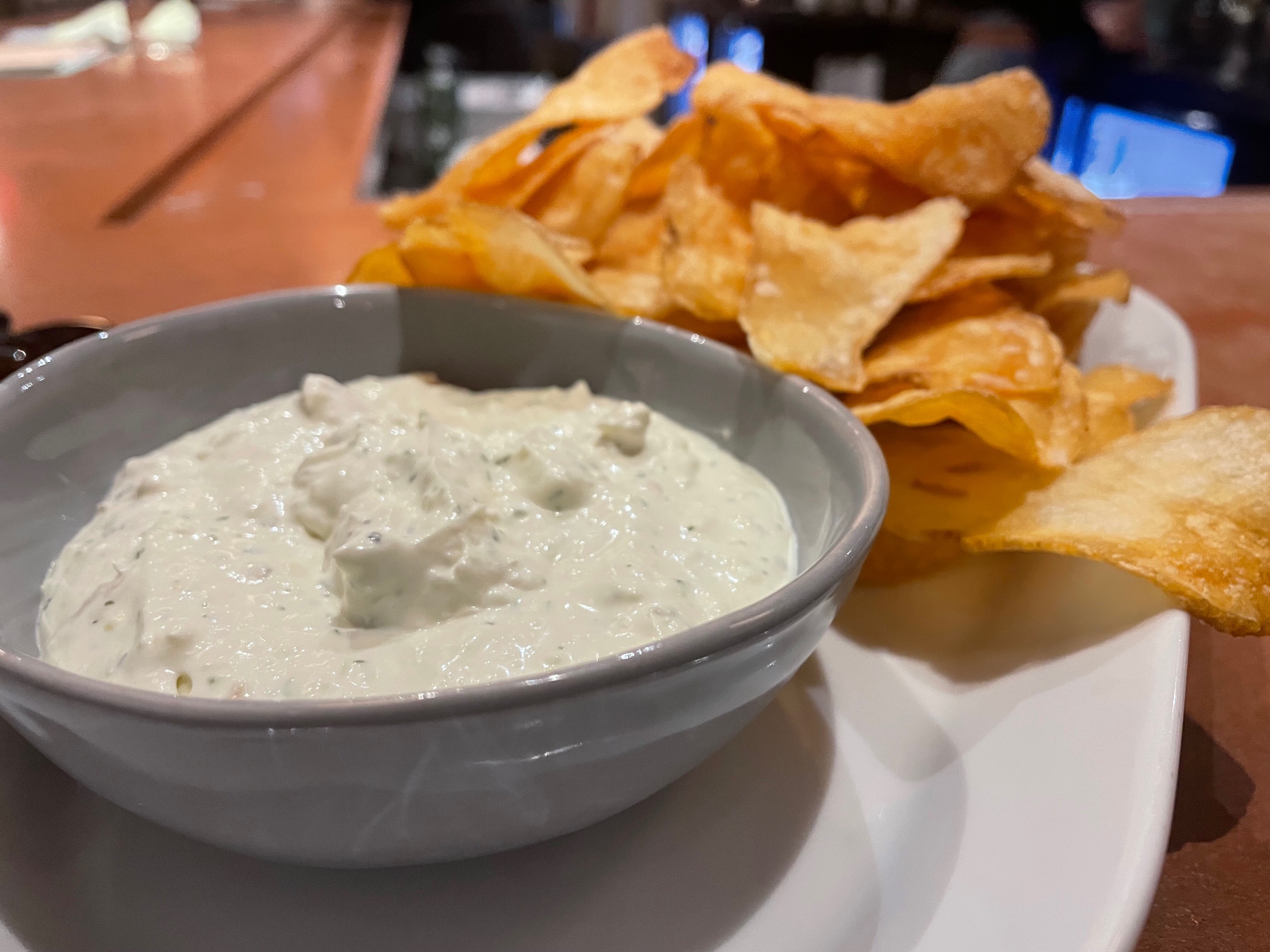 On a brown table, there is a long white platter with a bowl of white trout dip and a big portion of potato crisps. Photo by Alyssa Buckley.