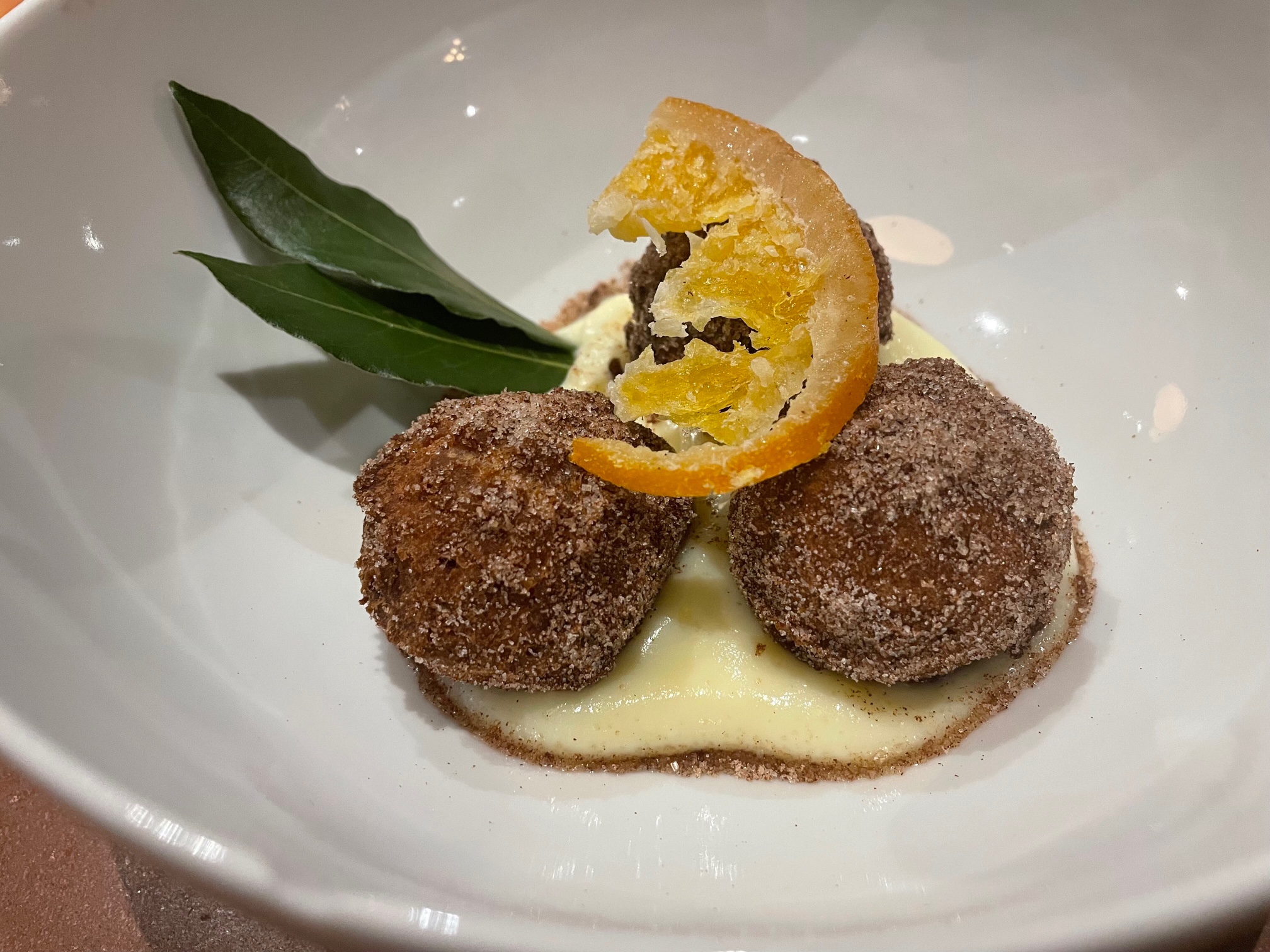 In a large white bowl, there are three balls of donuts covered in cinnamon sugar topped with a candied orange slice. Beneath the balls is a white pastry cream and two bay leaves. Photo by Alyssa Buckley.