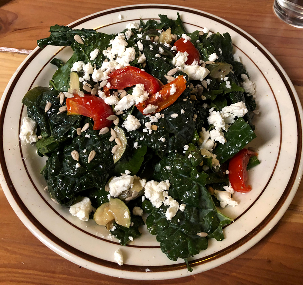 The kale bounty salad at Watson's in Champaign, Illinios. Tuscan kale salad with roasted vegetables, sunflower seeds, and goat cheese is served on a round white plate with a brown decorative edge. The plate is on a wood table. Photo by Jessica Hammie. 