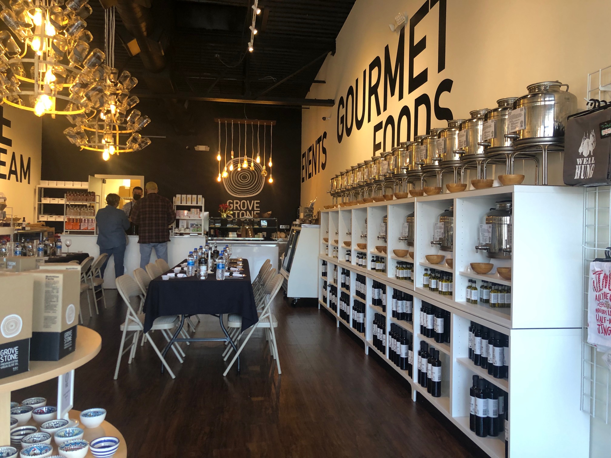 Inside Grovestone, there are white shelves on both left and right sides of the room filled with bottles of olive oil and vinegar. There is a long rectangular table with a black tablecloth and place settings. There are two customers checking out and a male masked cashier facing the camera. Photo by Alyssa Buckley.