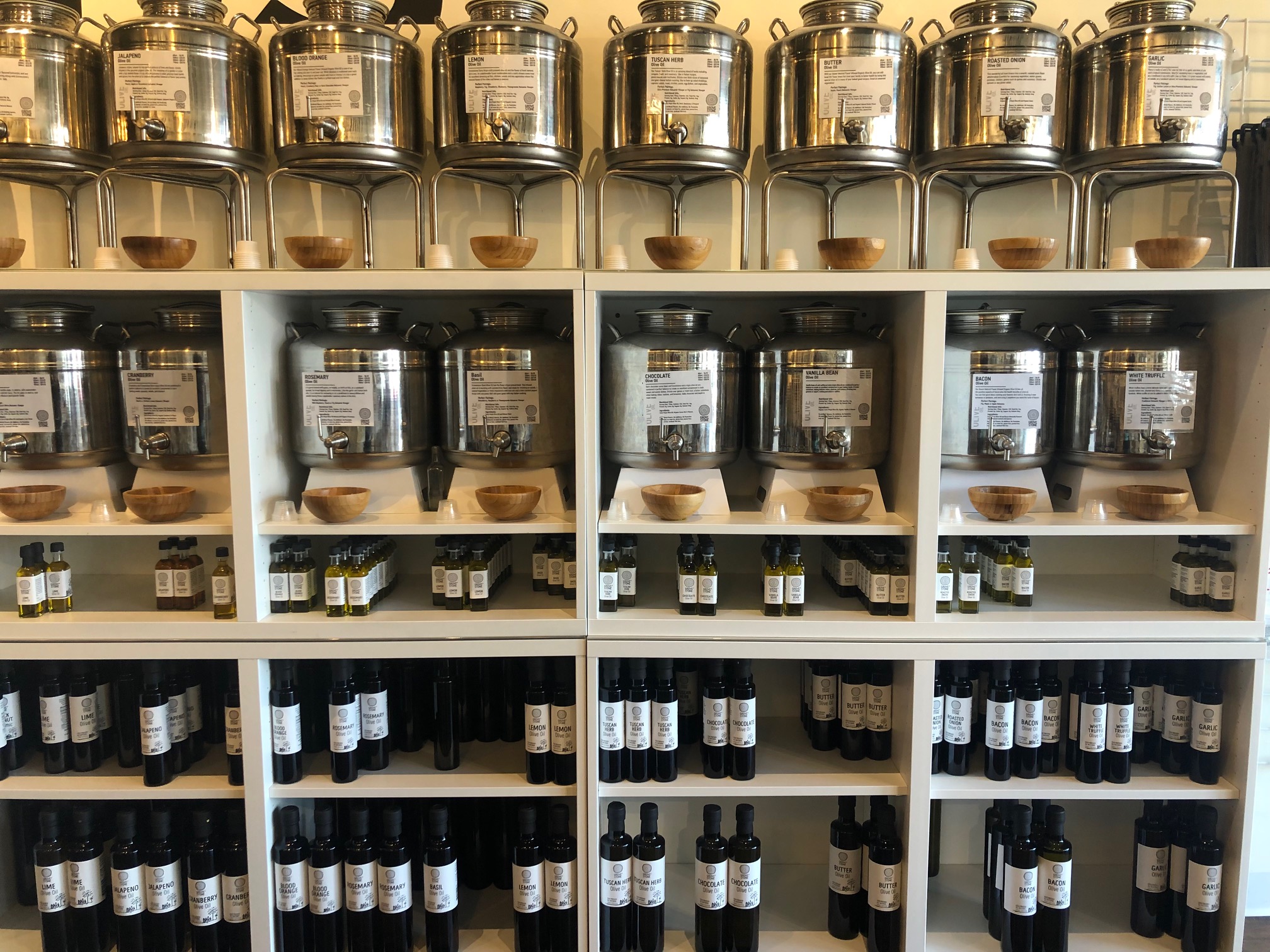 The white shelves are topped with silver beverage dispensers filled with infused olive oils. Below, the shelves have three sizes of bottles (small, medium, and large) on the shelf for sale. Photo by Alyssa Buckley.