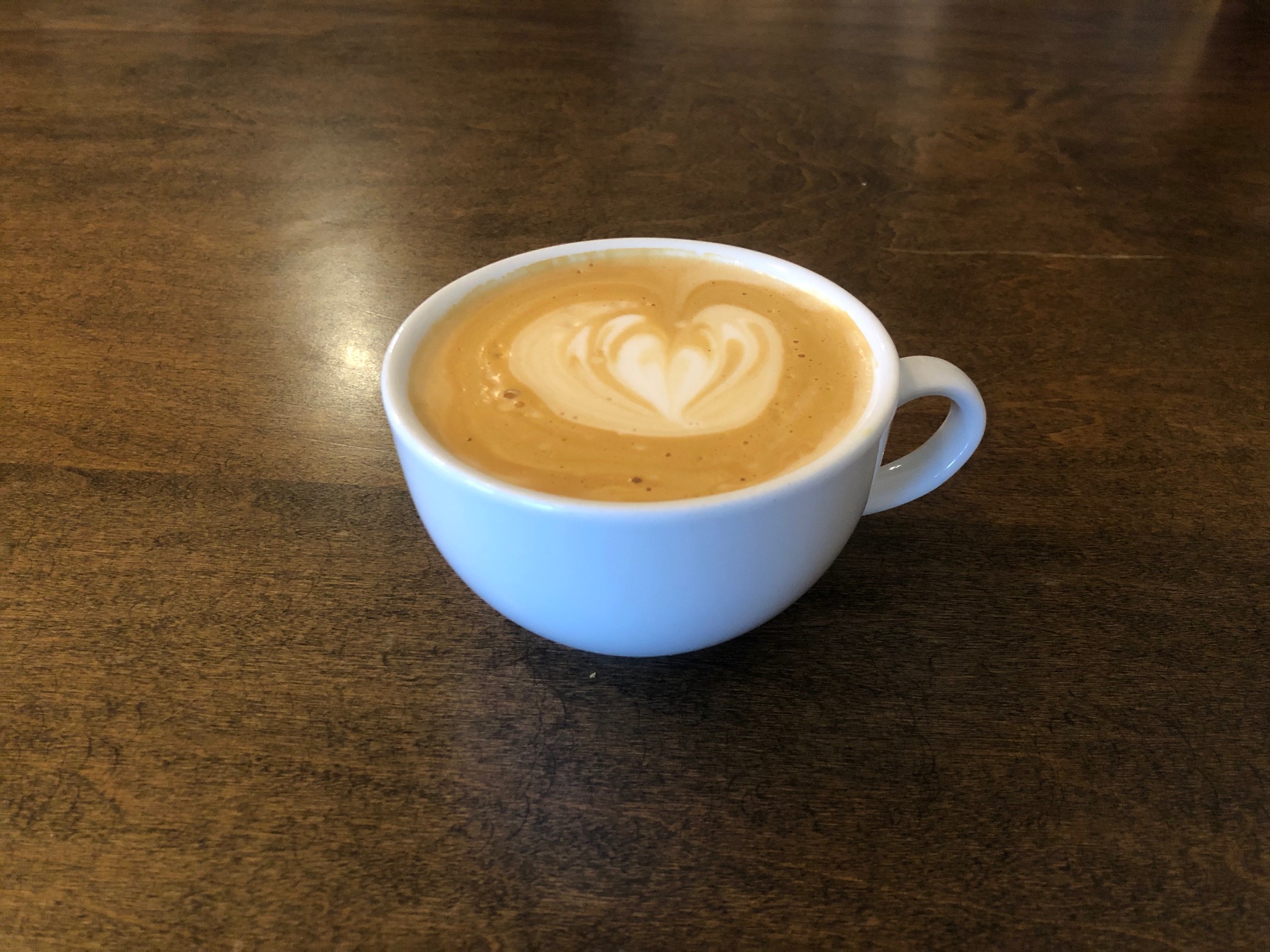 On a dark brown table, there is a white mug of coffee with a heart latte art in the foam. Photo by Alyssa Buckley.