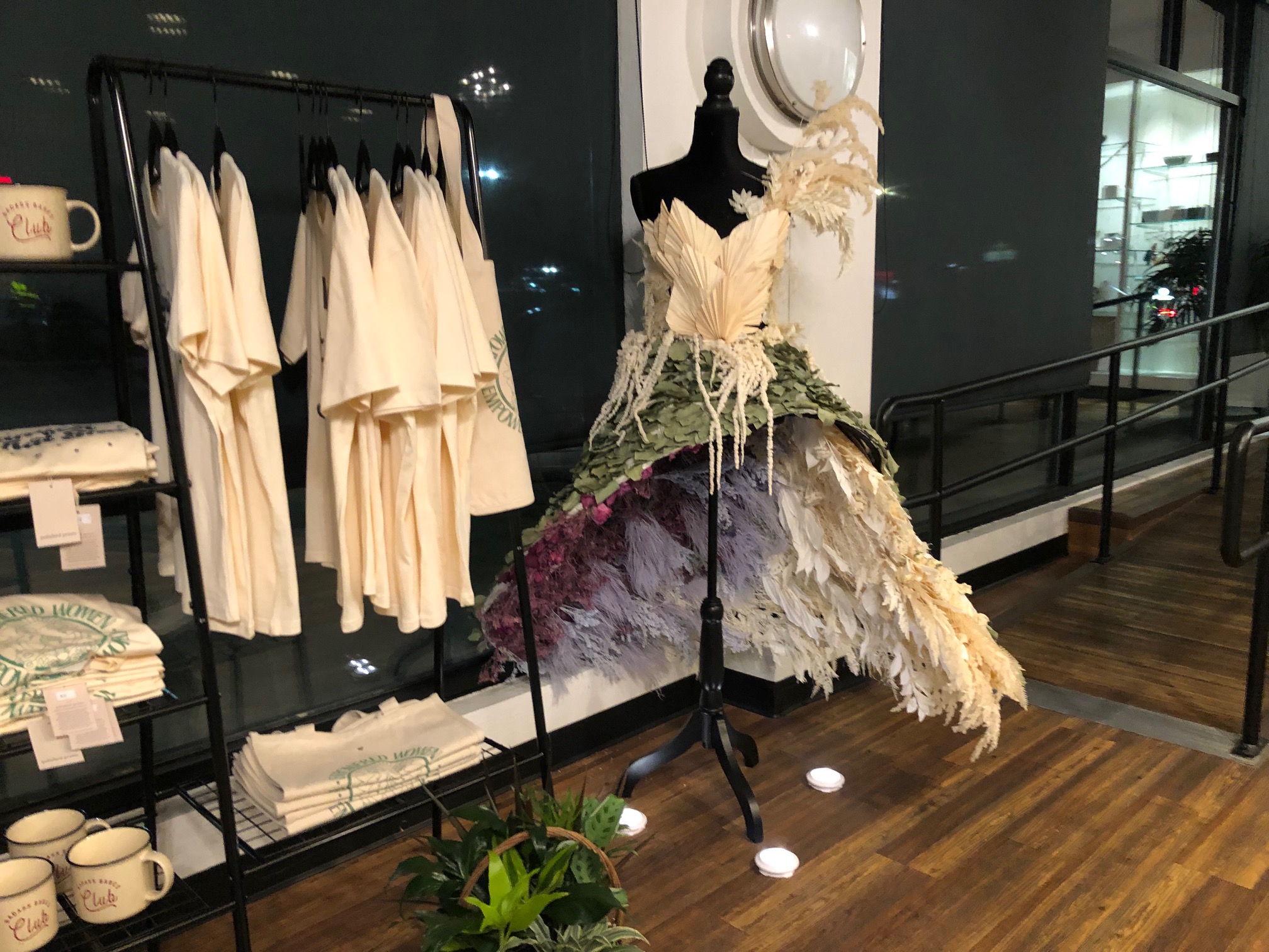 There is a beautiful white gown with feathers on a black mannequin, and several shirts on hangers in front of a black wall. Photo by Alyssa Buckley. 