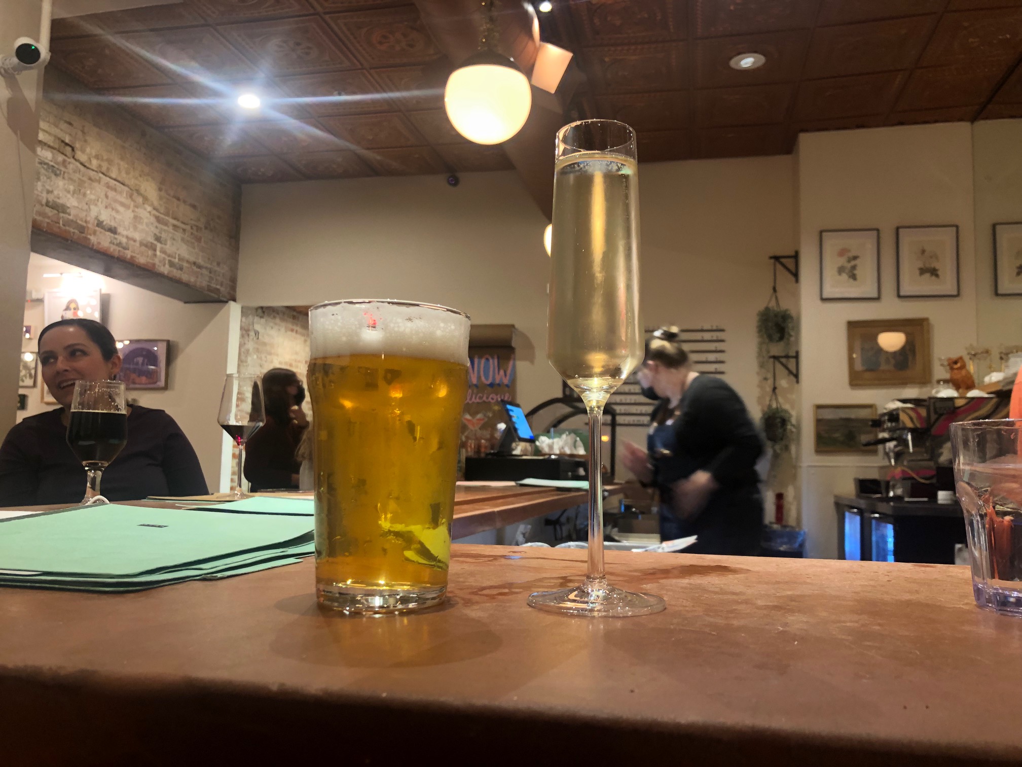 On a brown counter, there are two glasses: a beer glass with a light colored beer and a clear prosecco with tiny bubbles in a champagne flute glass.  Photo by Alyssa Buckley.