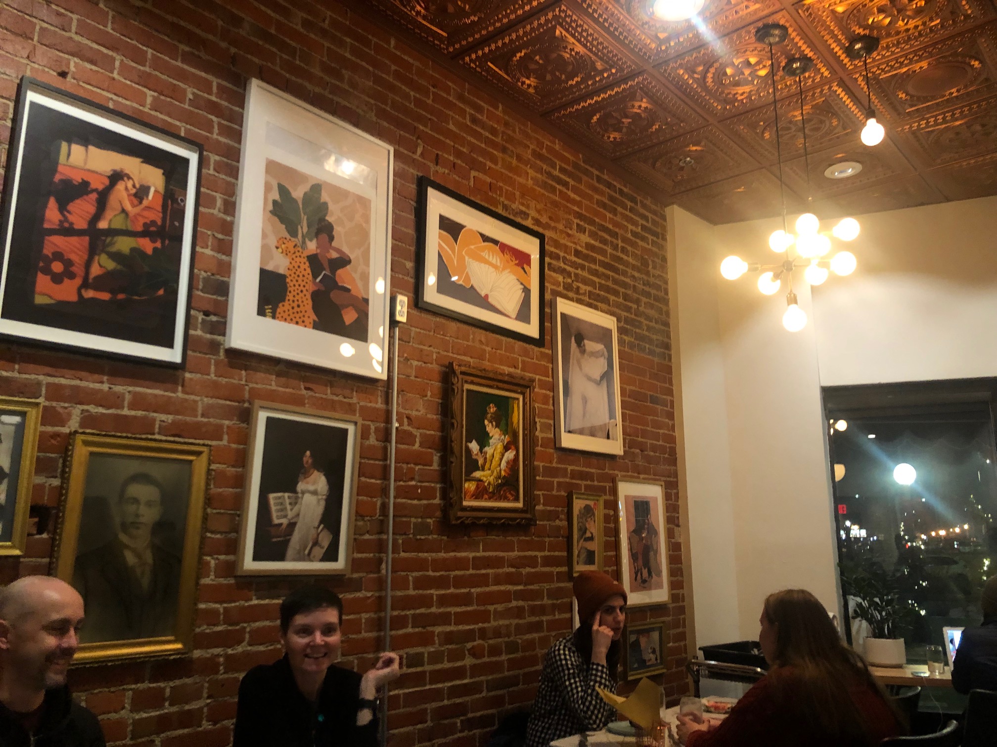 Inside The Literary, there are lots of framed artwork in a gallery wall on a brick wall. There are diners, unmasked, smiling on a Thursday evening.  Photo by Alyssa Buckley.