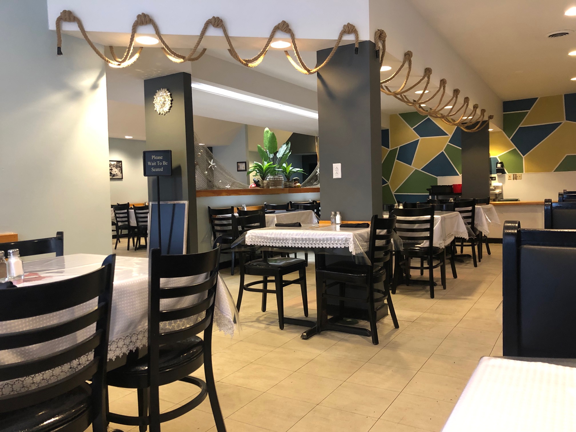 Inside La Bahia Grill, there are empty tables with white table cloths and black metal chairs. There is a feature wall with brightly colored interlocking shapes and nautical themes. Photo by Alyssa Buckley.