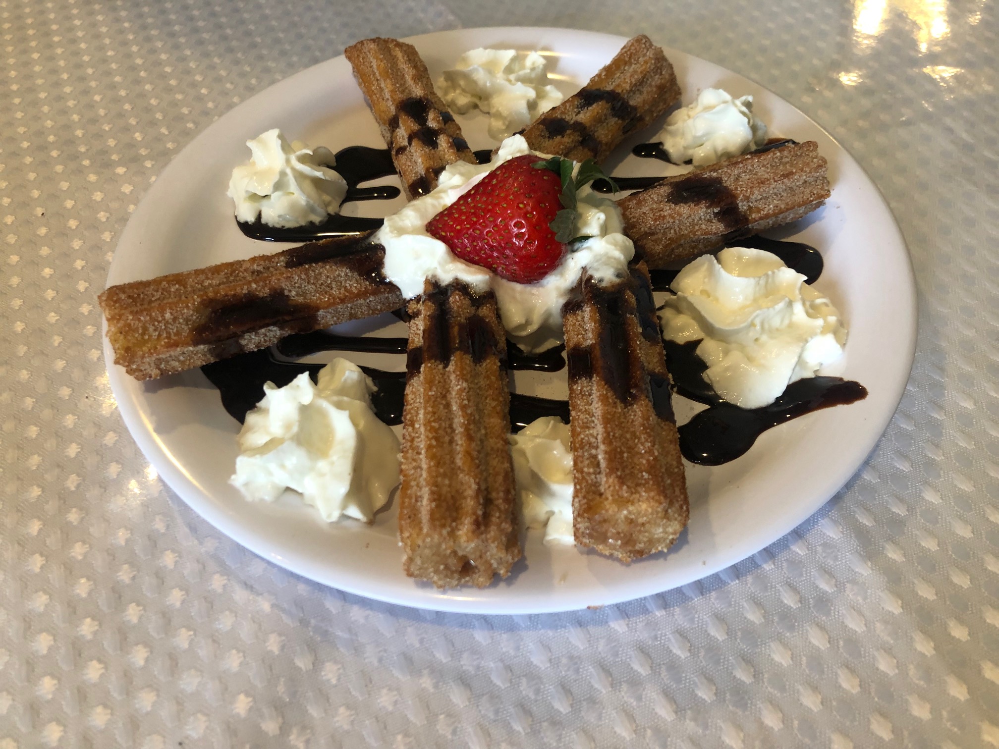 On a white plate, there are six churro sticks with chocolate syrup on top with whipped cream poufs. Photo by Alyssa Buckley.