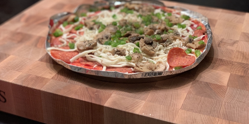 On a checkered butcher block kitchen island, there is a takeout pizza in a tin pan. The pizza is uncooked with shredded white cheese, pepperoni, sausage, and green peppers on top. Photo by Stephanie Wheatley.