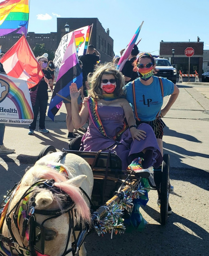 A woman is wearing a purple strapless gown, long brown wig, mask with big red lips, and a rainbow sash that says Grand Marshal. She sits in a carriage pulled by a miniature horse. Crouching next to her a is a person in a blue t-shirt, rainbow suspenders, and rainbow mask. They are both wearing sunglasses. Photo from UP Center Facebook page.