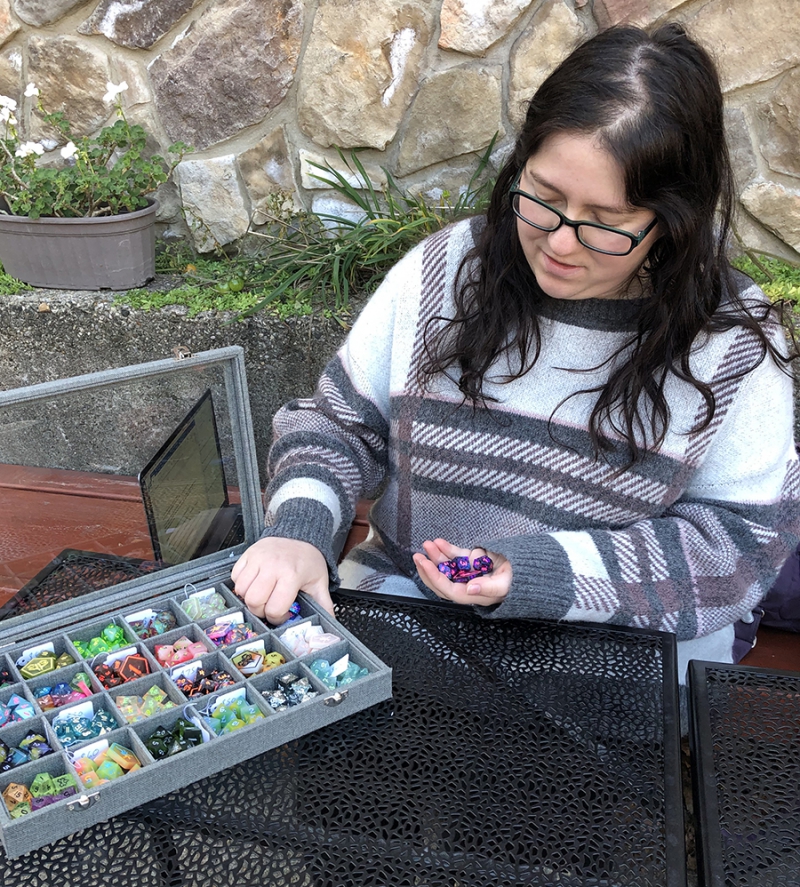 A woman with long dark hair and black rimmed glasses is sitting at a wrought iron table. She is wearing a black, gray, and white plaid sweater, and scooping dice from the container. Photo by Cope Cumpston. 