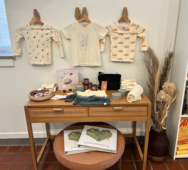 Three child outfits hang on the wall above a small wooden table with other items for children. Underneath the table there is a stool with two photo albums stacked on it. Photo by Dani Nutting.