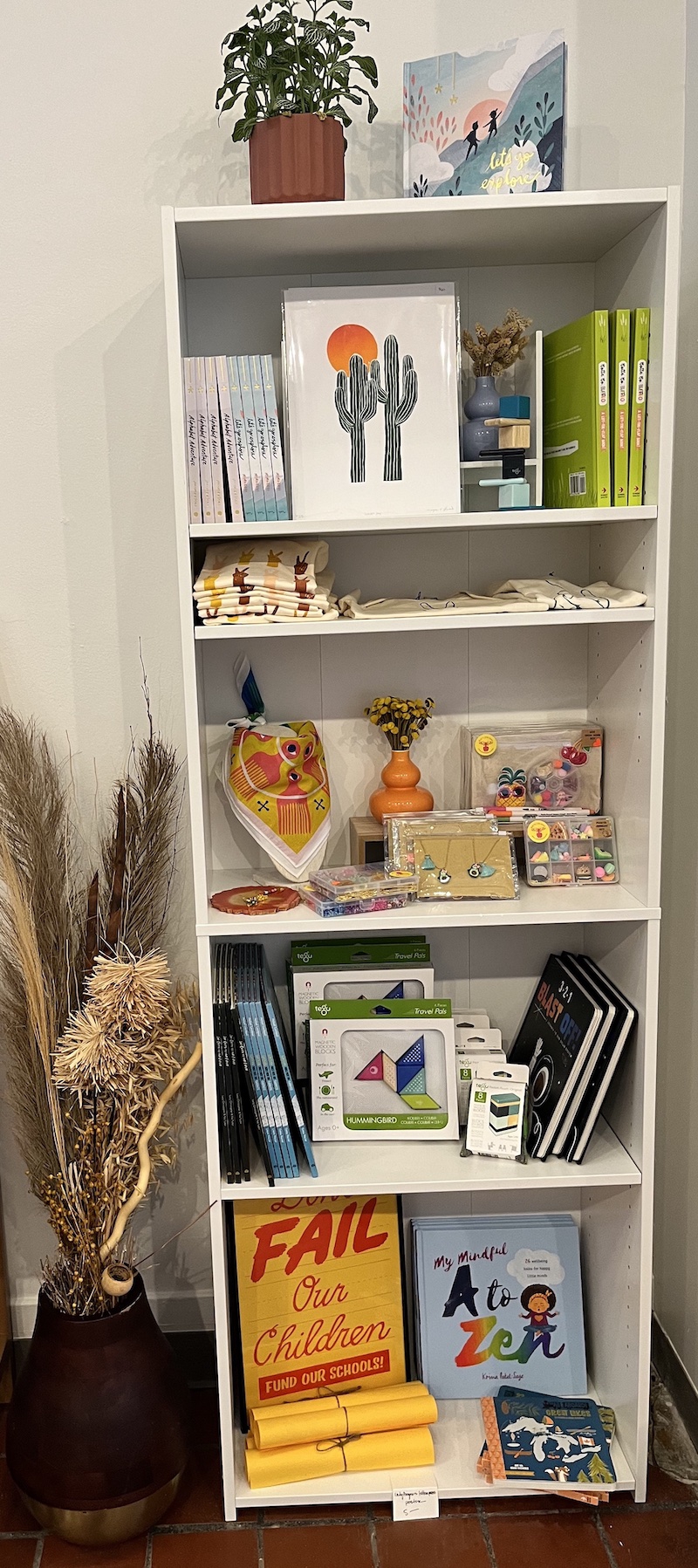 A tall white shelving unit displaying children's items: books, folded clothing, jewelry, and games. A potted plant sits on top. Photo by Dani Nutting. 