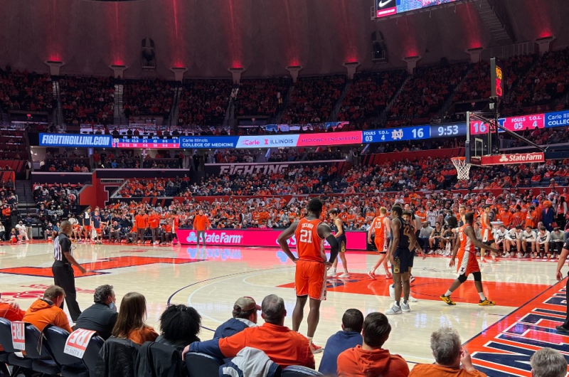 The basketball court at State Farm Center. Several players are standing on the court, the closest player has orange shorts and jersey with the number 21 and Cockburn in white. People are filling the stands around the perimeter. Photo by Julie McClure.