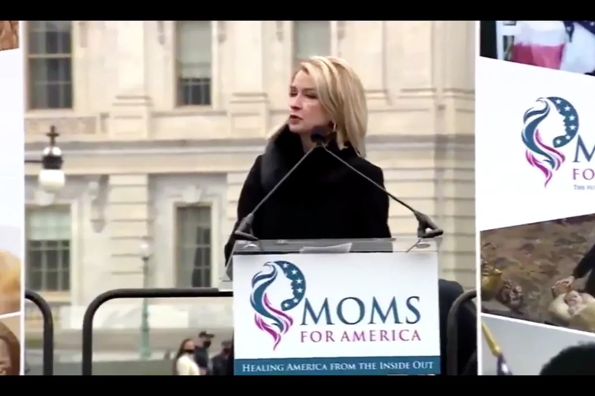 Screenshot of video of Representative Mary Miller (IL-15) speaking outside at a podium. She is a thin, blonde white woman wearing a black coat with a fur collar. The lecturn has two microphones pointed at the center of the space. There is a sign on the front of the lecturn that says 