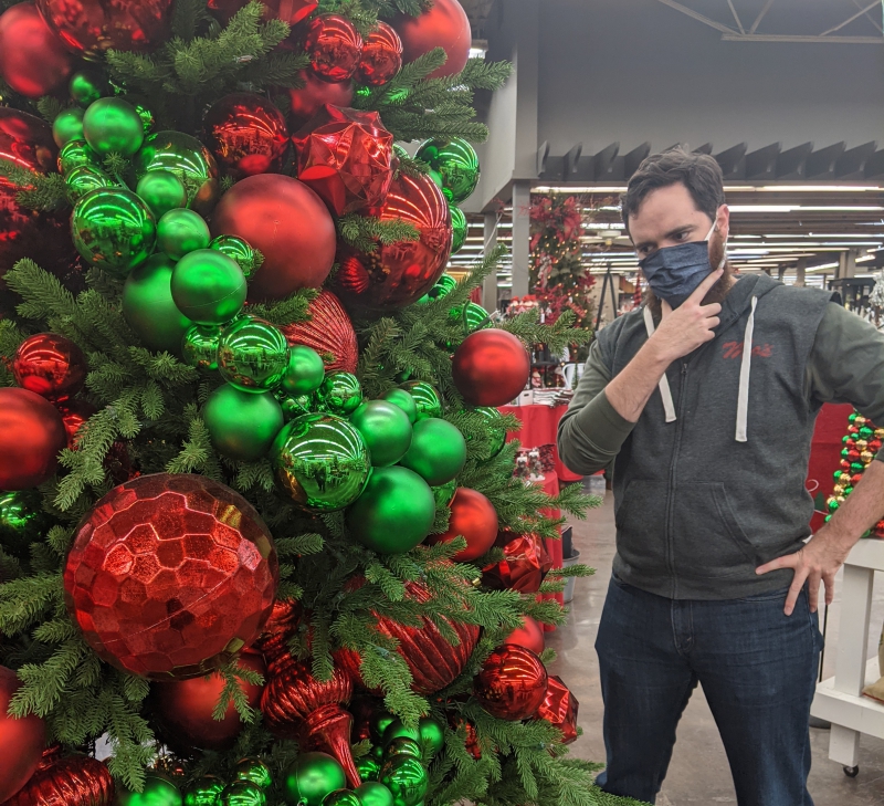 The writer is looking at a Christmas tree covered in varying sizes of red and green ball ornaments. He has his hand on his chin like he is contemplating something. Photo by Andrea Black.
