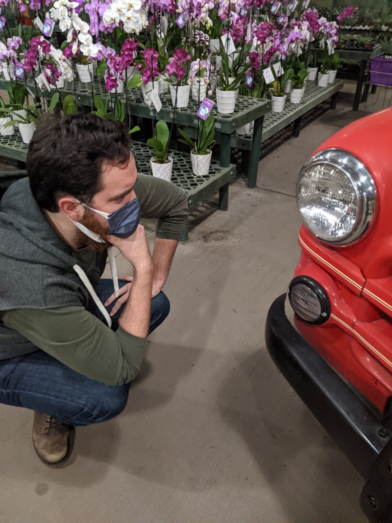 The writer is crouched down staring into the headlight of the front of a red car. Behind him are platforms with potted flowers. Photo by Andrea Black. 
