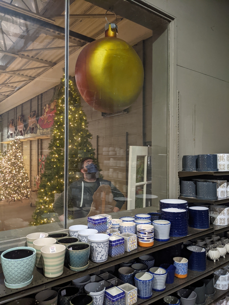 A view of the writer through a large window. He is staring up at a large gold circle hanging from the ceiling. There are lighted Christmas trees behind him and rows of pottery on shelves below the window. . Photo by Andrea Black.