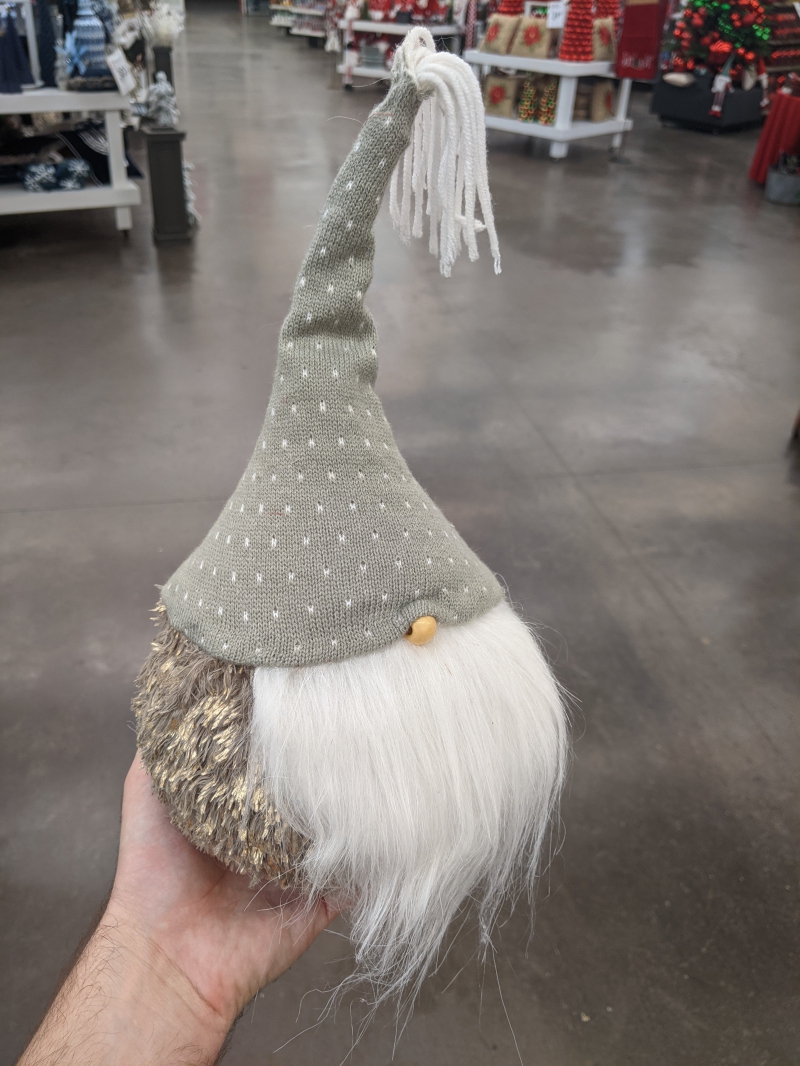 A round fuzzy gnome decoration with a long white beard, tiny round nose, and gray knit hat. It is being held by the writer. Photo by Tom Ackerman.