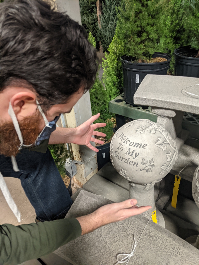 The writer is leaning over looking at a cement orb that says Welcome to My Garden. His hands are outstretched towards it. Photo by Andrea Black. 