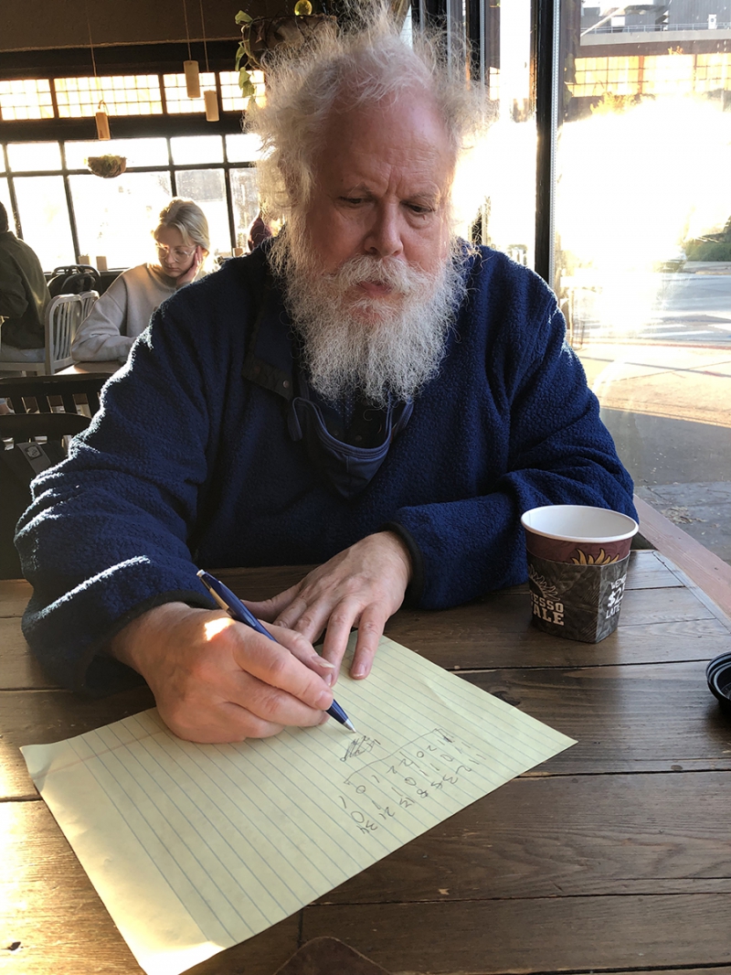 A man with white hair and a long beard is sitting at a wooden table. He is wearing a blue shirt and is writing numbers on a piece of legal paper. Photo by Cope Cumpston. 