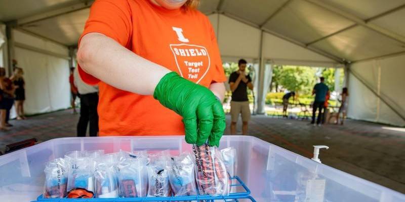 A woman in an orange shirt with black gloves is putting vials in a plastic tub. Photo from Illini Union Facebook page.