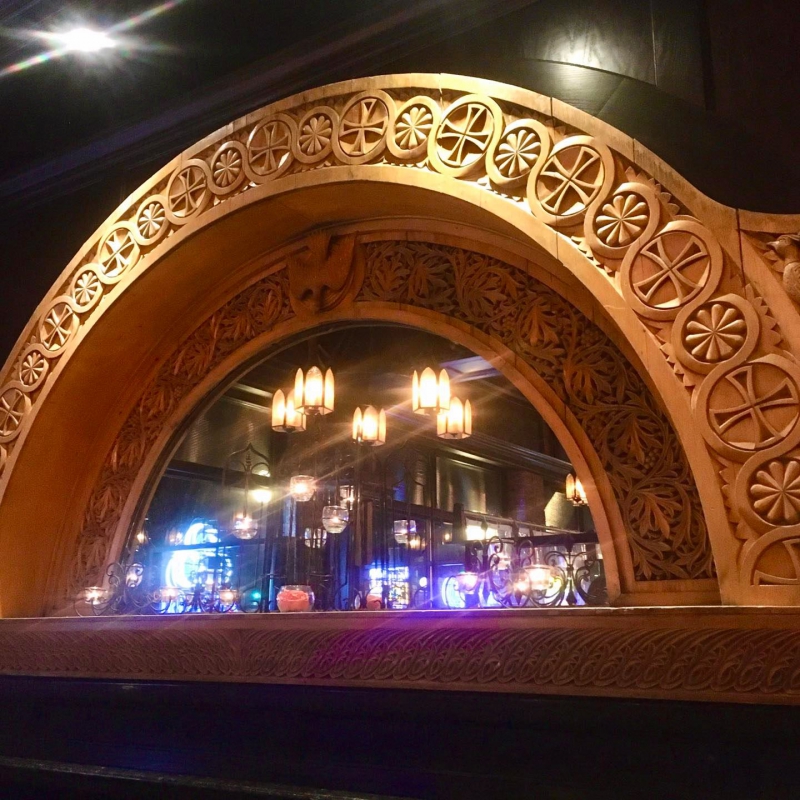 A semi-circle mirror framed by ornate wood carved arch. There are hanging lights reflected in the mirror. Photo from Seven Saints Facebook page.
