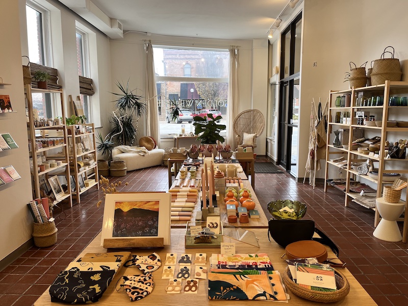 The interior of a store. In the center is a long wooden table filled with merchandise: earrings, zipper pouches, candles, stemmed glasses. There are shelves along the walls with more items, and a window out to the street in the background. Photo by Dani Nutting.
