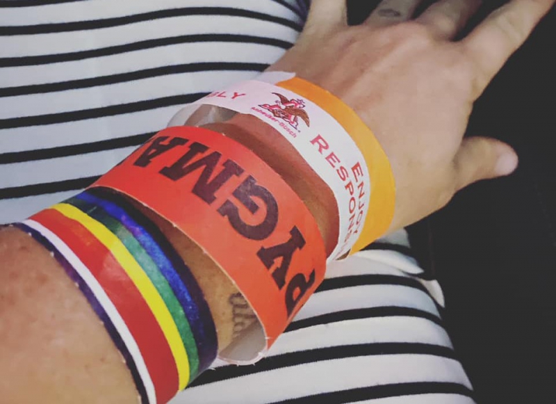 Close up of a wrist with several paper wristbands: one is rainbow, one is red with PYGMALION in black letters, one is white and says Enjoy Responsibly in white, one is solid orange. Photo by Julie McClure.