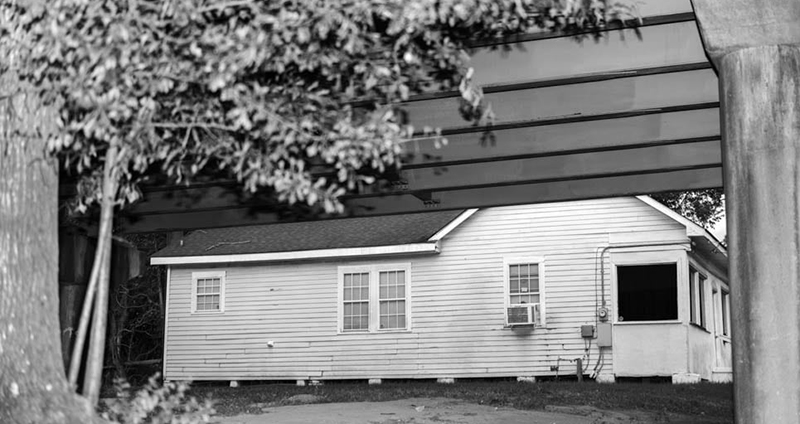 Black and white photo of a house below a tree and blocked off by bridge.